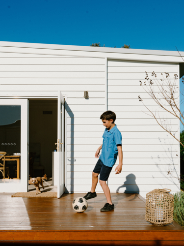 Young boy practicing soccer on sunlit deck outside home - Australian Stock Image
