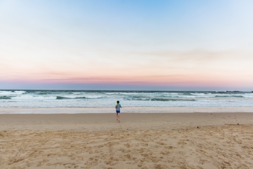 Young boy running from sand towards waves on beach at sunset