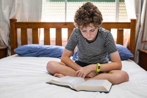 Young boy sitting cross legged on bed reading a book natural light - Australian Stock Image