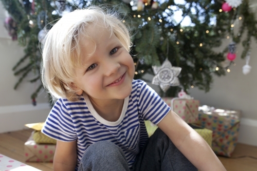 Young boy sitting in front of Christmas tree smiling - Australian Stock Image