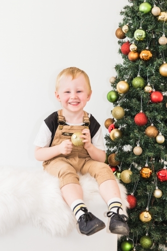 Young boy sitting next to Christmas tree smiling holding bauble decoration - Australian Stock Image