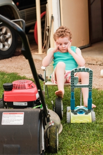 Young boy sitting on chair resting head with foot on lawn mower - Australian Stock Image