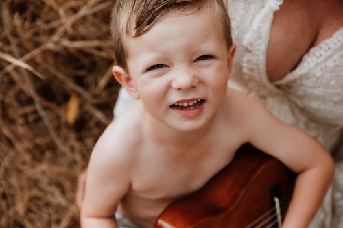Young boy sitting on mother's lap in long grass, smiling up at camera - Australian Stock Image