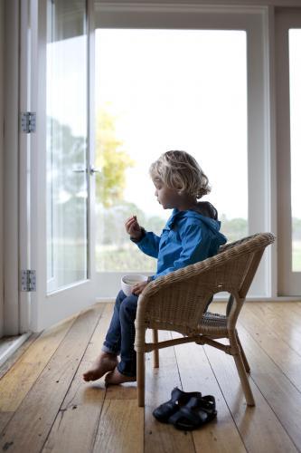 Young boy sitting on wicker chair inside, eating a snack - Australian Stock Image