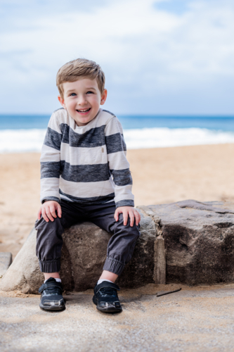 Young boy smiles while sitting on a rock near the beach with ocean in the background - Australian Stock Image