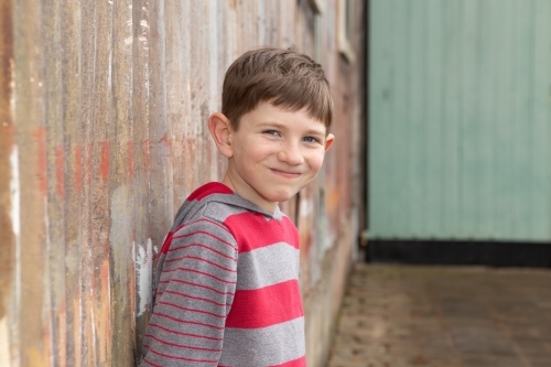 Young boy standing in front of rusty wall smiling - Australian Stock Image