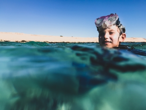 Young boy swimming in ocean with snorkelling mask on head looking at camera smiling