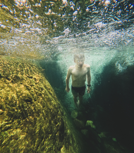 Young boy underwater surrounded by bubbles in clear dark green waters, rocks in background - Australian Stock Image