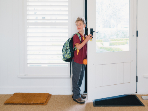 Young boy wearing backpack standing at the doorway holding a small orange ball - Australian Stock Image