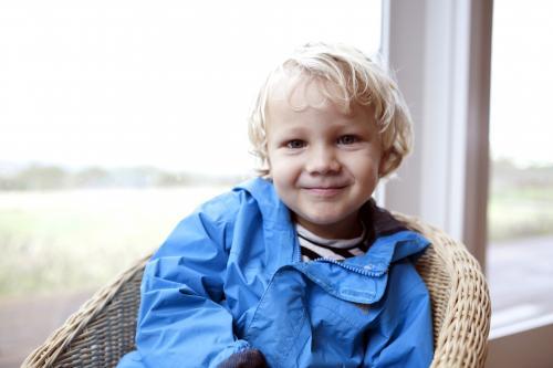 Young boy wearing blue jacket sitting in wicker chair in front of a window - Australian Stock Image