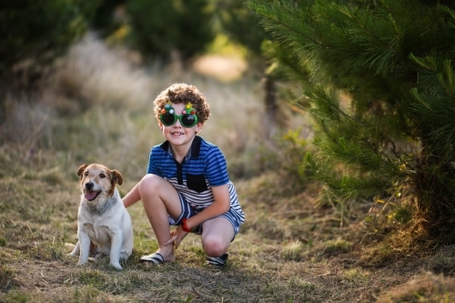 Young boy wearing glasses squatting next to dog in pine trees - Australian Stock Image