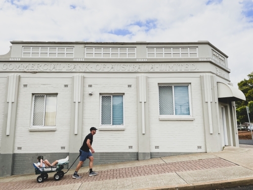 Young boy with broken leg being wheeled in cart by man walking past historical building - Australian Stock Image