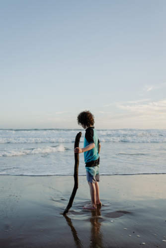 Young boy with curly hair holding a large stick standing on a beach in the afternoon - Australian Stock Image
