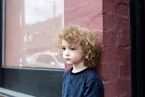 Young boy with curly hair standing against a pink brick wall
