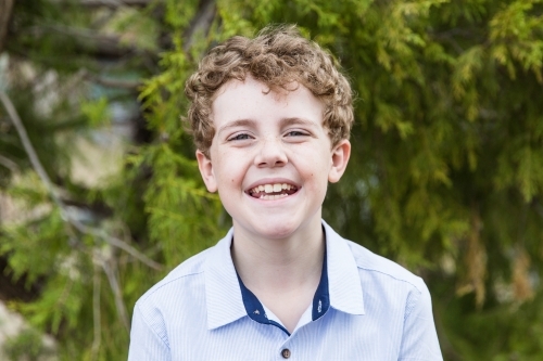 Young boy with curly hair standing in front of tree smiling