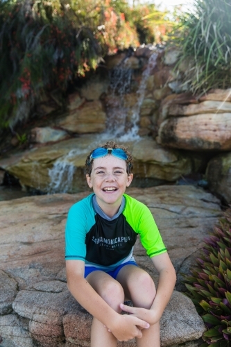 Young boy with goggles sitting on rocks in front of waterfall happy smiling - Australian Stock Image