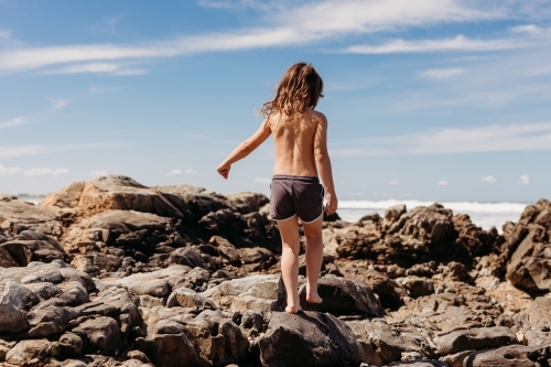 Young boy with long hair walking on rocks in ocean