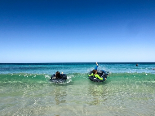 young boys in wetsuits lying on inflatable tyres on small beach waves - Australian Stock Image