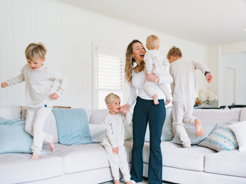 Young boys playing and jumping on the couch with mum carrying the baby - Australian Stock Image