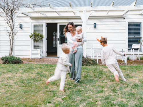 Young boys running around mum holding a baby in the garden - Australian Stock Image
