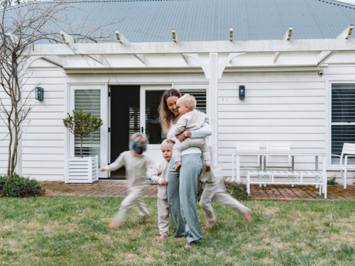 Young boys running around mum holding a baby in the garden - Australian Stock Image
