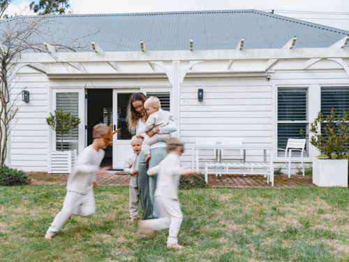 Young boys running around mum holding a baby in the garden - Australian Stock Image