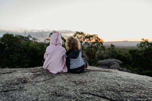 Young brother and sister sitting together on a rocky ledge in the Australian bush watching a sunset