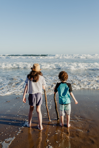 Young brother & sister (young boy and girl) holding sticks standing together at the beach - Australian Stock Image