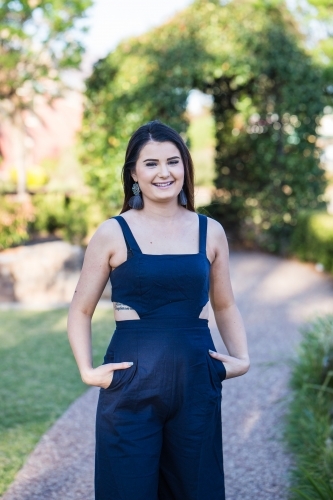 Young brunette woman with tattoo standing in garden smiling with hands in pockets - Australian Stock Image