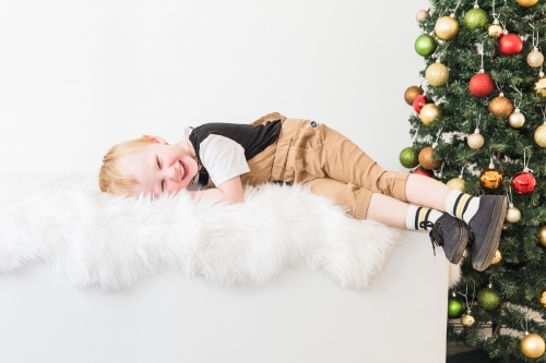 Young child lying on white rug laughing with Christmas tree - Australian Stock Image