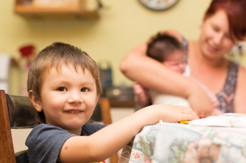 Young child sitting at table with mother and baby in background - Australian Stock Image