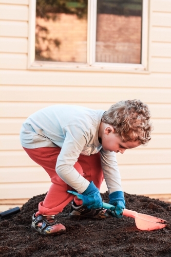 Young child standing on pile of dirt wearing gloves working with shovel - Australian Stock Image