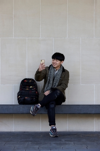 Young Chinese man sitting on bench using mobile phone - Australian Stock Image