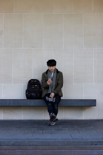 Young Chinese man sitting on bench using mobile phone - Australian Stock Image