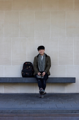 Young Chinese student sitting on bench at university campus with mobile phone