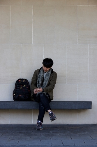 Young Chinese student sitting on bench at university campus with mobile phone - Australian Stock Image