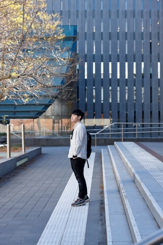 Young Chinese student standing on stairs smiling with buildings and tree behind - Australian Stock Image