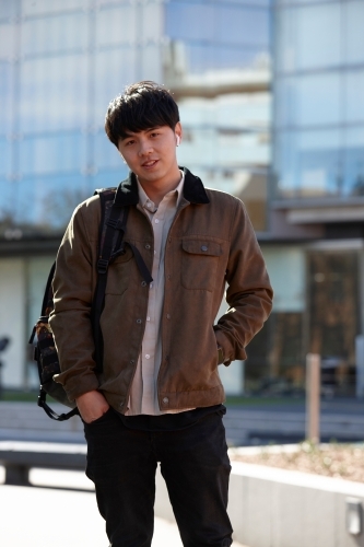 Young Chinese student standing with hands in pocket with listening to wireless headphones - Australian Stock Image