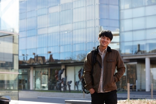 Young Chinese student standing with hands in pocket with listening to wireless headphones - Australian Stock Image