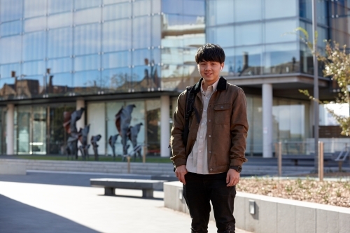 Young Chinese student standing with hands in pocket with listening to wireless headphones - Australian Stock Image