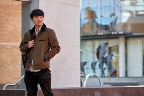 Young Chinese university student standing with hands in pocket listening to wireless headphones - Australian Stock Image