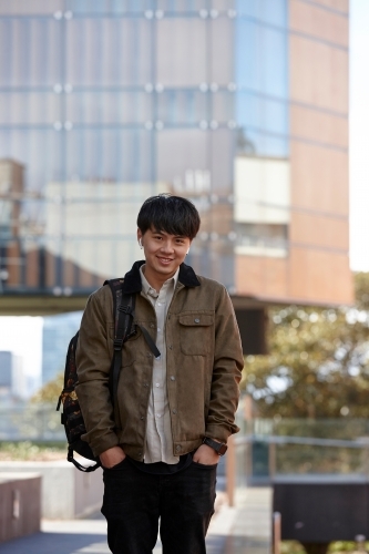 Young Chinese university student standing with hands in pocket listening to wireless headphones - Australian Stock Image