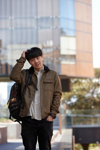 Young Chinese university student standing with hands in pocket listening to wireless headphones - Australian Stock Image