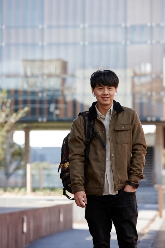 Young Chinese university student standing with hands in pocket listening to wireless headphones - Australian Stock Image