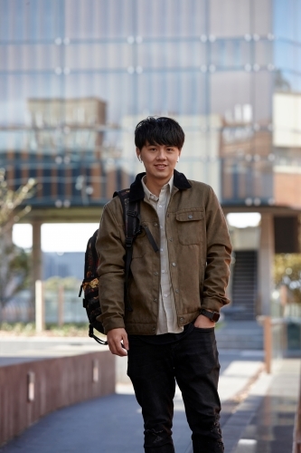 Young Chinese university student standing with hands in pocket listening to wireless headphones - Australian Stock Image