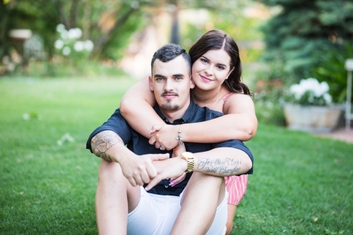 Young couple sitting on grass in garden with woman's arms around man's neck smiling - Australian Stock Image
