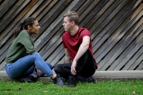 Young couple sitting on the ground talking in front of textured wooden panels - Australian Stock Image