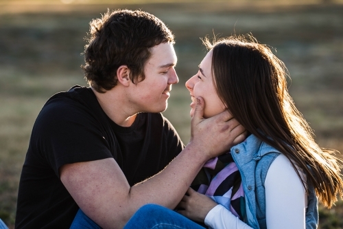 Young couple sitting together laughing with man holding girlfriend's face - Australian Stock Image