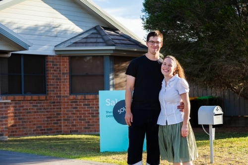 Young couple stand in front of sold for sale sign on first home - Australian Stock Image