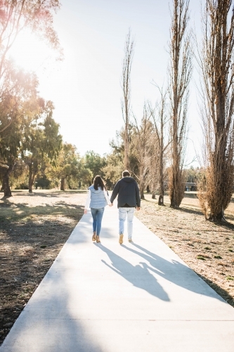 Young couple walking away into distance on path in park holding hands - Australian Stock Image